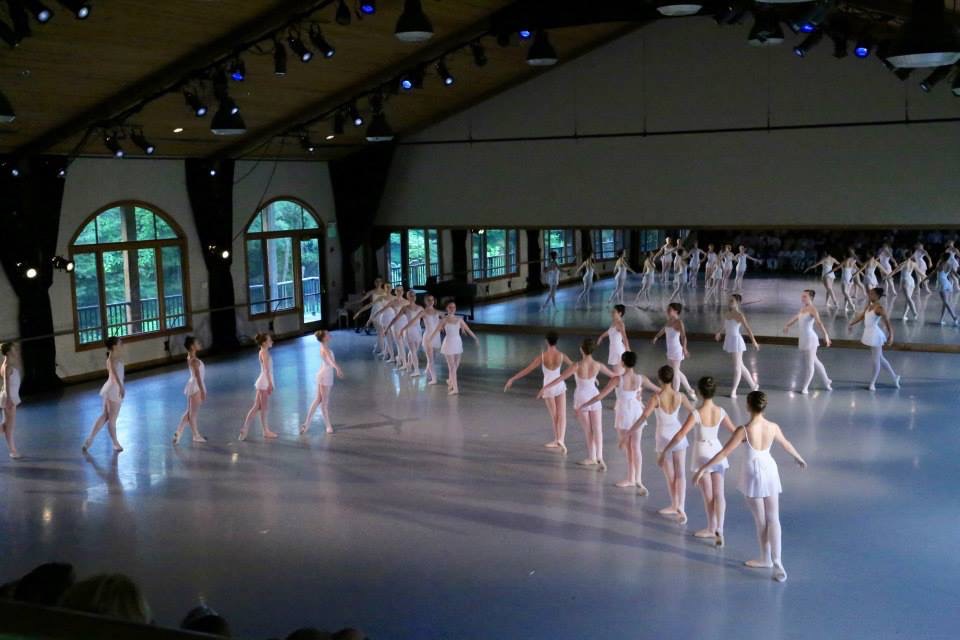 In a large, airy dance studio, four lines of teenage female dance students stand in B+, facing in towards center and creating a pinwheel formation. They wear white leotards and skirts, pink tights and ballet slippers and all wear their hair in buns. Stage lighting from above lights the dancers.