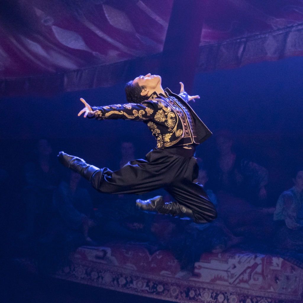 During an onstage performance, Jeffrey CIrio jumps high in the air with his legs bent behind him, his upper body arched back and his arms spread wide. He wears black pants and boots and a short black jacket with gold brocade.