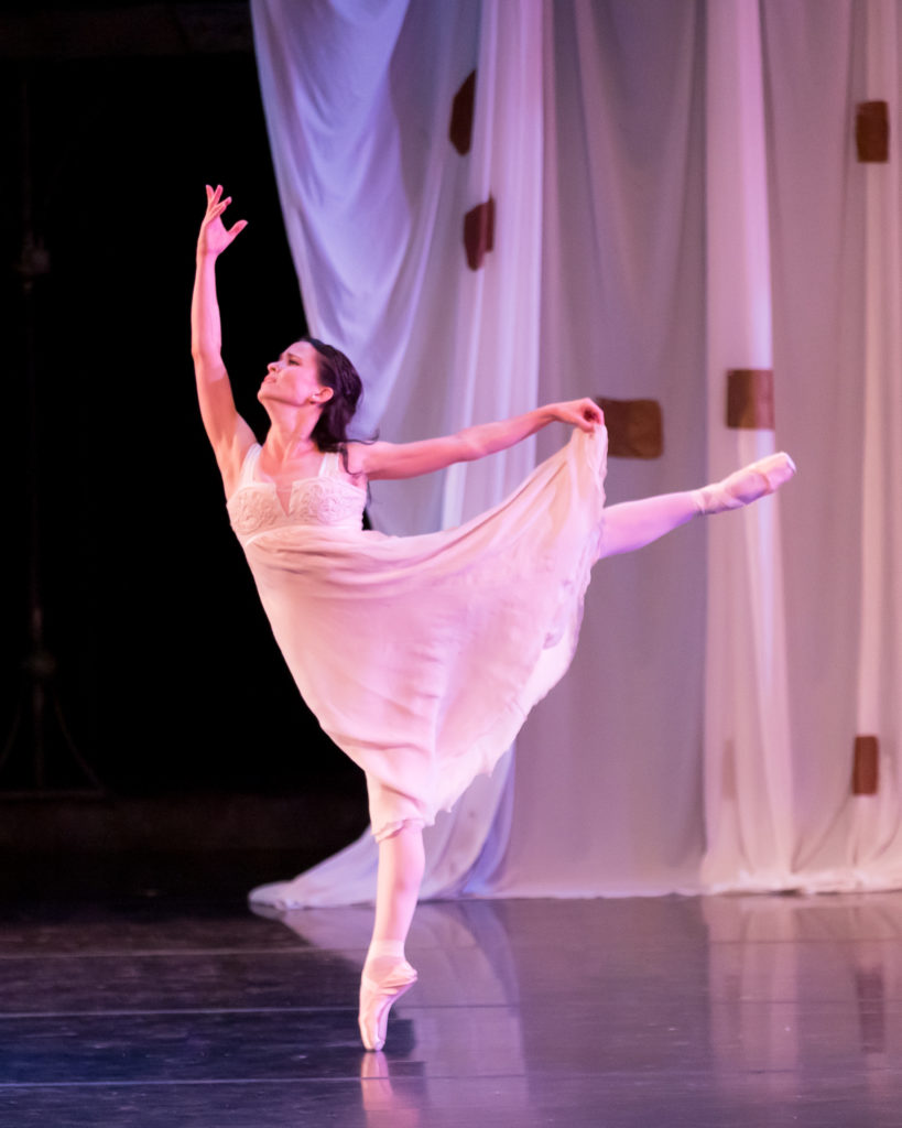 Mollie Sansone dances onstage during a performance, wearing a long pikn nightgown, pink tights and pointe shoes. She does a piqué arabesque on pointe with her left leg up, holding her skirt with her elft hand and holding her right arm up above her head. A long white curtain hangs loosely in the background.