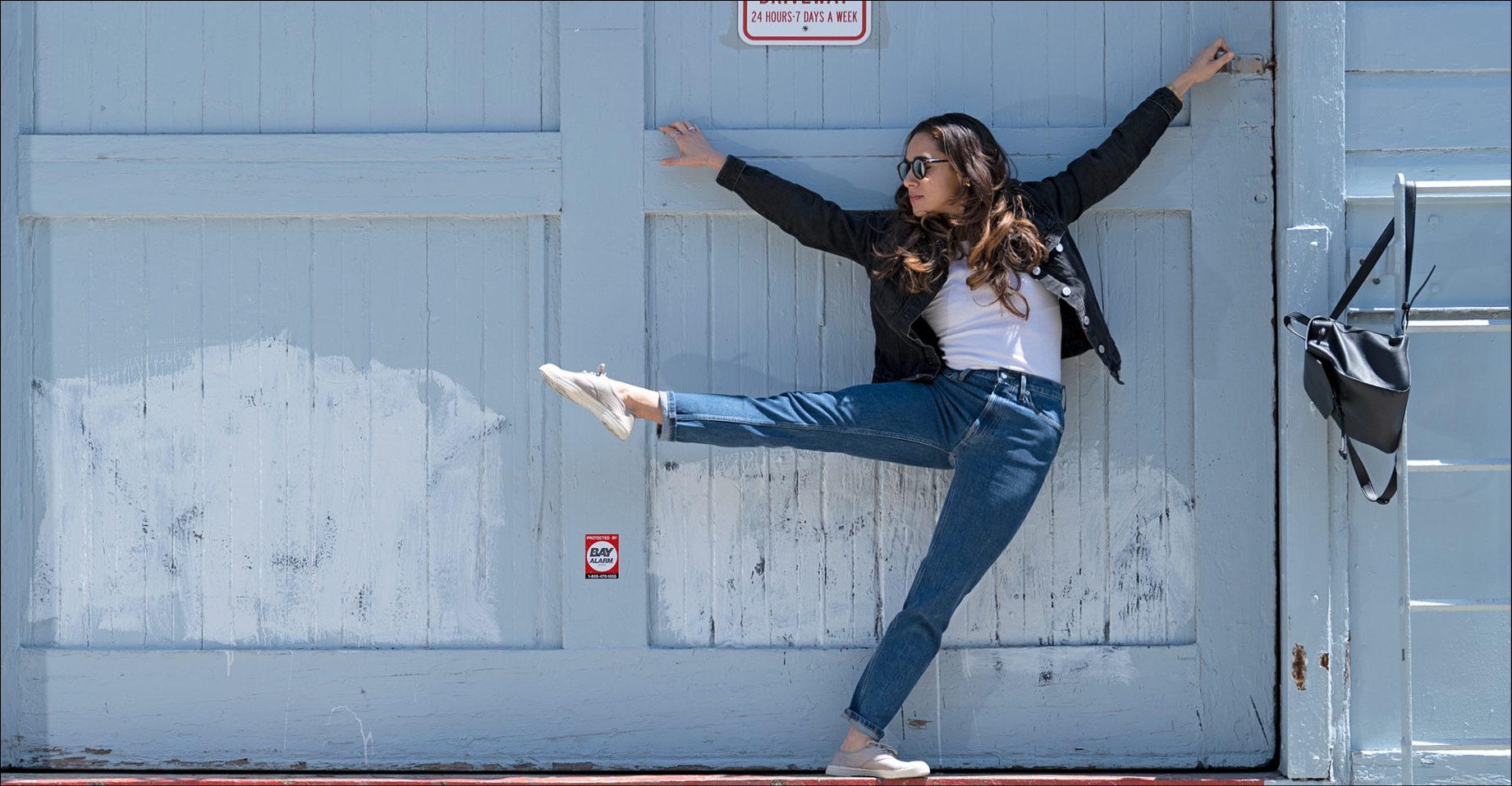 Tess Lane poses in a white t-shirt, black denim jacket, blue jeans, and light gray sneakers. She leans against a blue gray wooden plank wall holding onto the edge of a plank above her. Her arms are held upward on diagonals. She wears black, round sunglasses and her hair topples over her shoulders in waves as she looks to the right. Her torso leans slightly to the left to counterbalance her outstretched right leg, held a la seconde hip level with her foot pointed. Her left leg supports her on a diagonal as she leans off balance. A black leather backpack hangs on a metal bar to her left by a single strap.