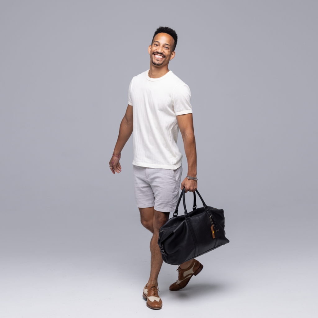 Ricardo Rhodes carries his black dance bag. He is wearing a cream-colored, short-sleeved shirt, gray shorts and brown-and-cream-colored Oxford shoes.