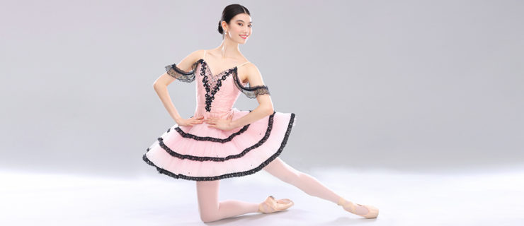 Chloe Misseldine, in a light pink pancake tutu with black lace embellishments and shoulder drapes, poses confidently with her left leg in tendu derriere, propped up on her right knee, her leg turned out. She wears light pink ballet tights and pointe shoes, and dainty white pearl drop earrings. With her hands on her hips and her head turned gently to the side, she smiles at the camera. Her dark hair is pulled back into a sleek low bun with a middle part, and she poses on a light gray backdrop.