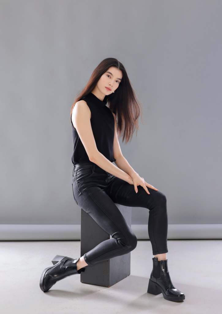CHloe Misseldine sits on a black, rectangular box with her body facing the right corner. She wears a black sleeveless shirt, black jeans and chunky black boots, and her hair is long and blowing slightly in a breeze. She sits with her hands crossed on her left thigh and her right leg bent back slightly, and looks toward the camera with a closed mouth smile.