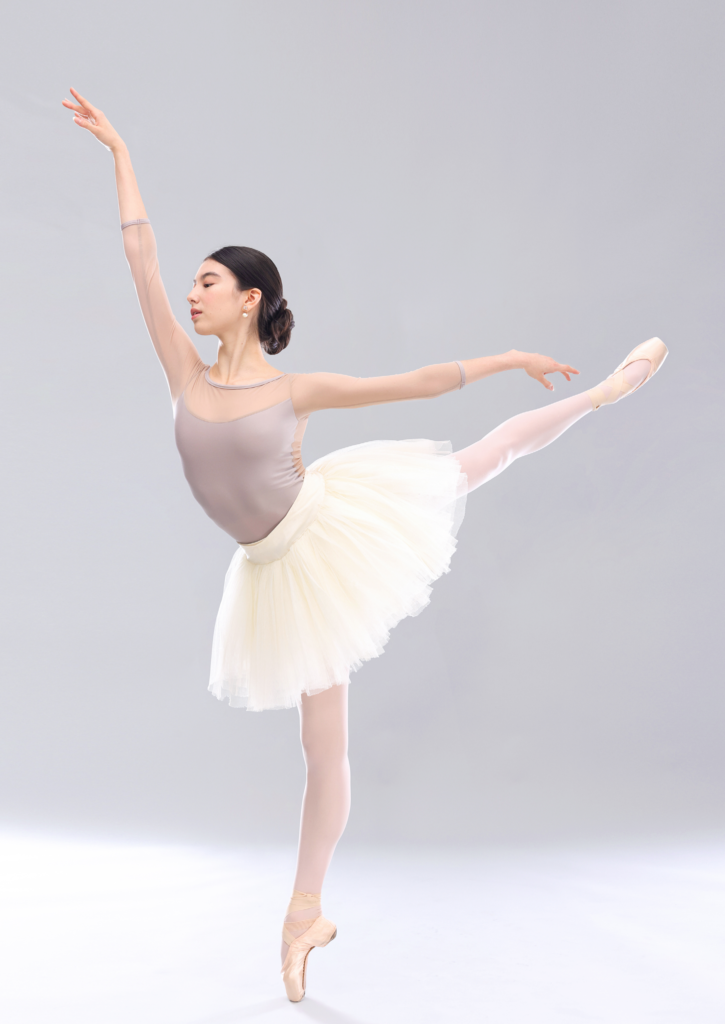 CHloe Misseldine poses in a first arabesque on pointe with her left leg behind her, and in front of a gray backdrop. She wears a beige leotard with mesh three-quarter sleeves, a white practice tutu, pink tights and pointe shoes. She looks down with her eyes and wears her hair in a low and full chignon.