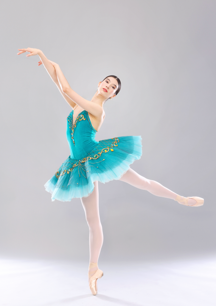 CHloe Misseldine poses in a low arabesque on pointe with her left leg behind her. She lifts her right arm up, drapes her left hand over her right forearm, and looks out towards the camera with a small smile. She wears a turquise pancake tutu with gold trim, pink tights and pointe shoes.