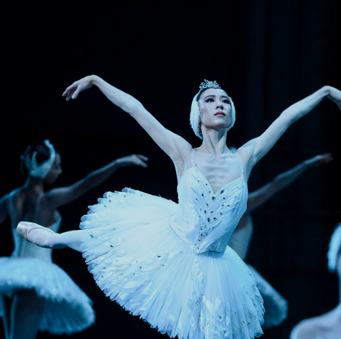 Sae Eun Park, costumed in a white tutu with feather embellishments, a white feathered headpiece with tiara, pink tights and pointe shoes, performs an arabesque with her right leg behind her. She lifts her arms up into a V-shape. Behind her, a corps of women in white tutus and feathered headpieces pose with their arms out to the side.