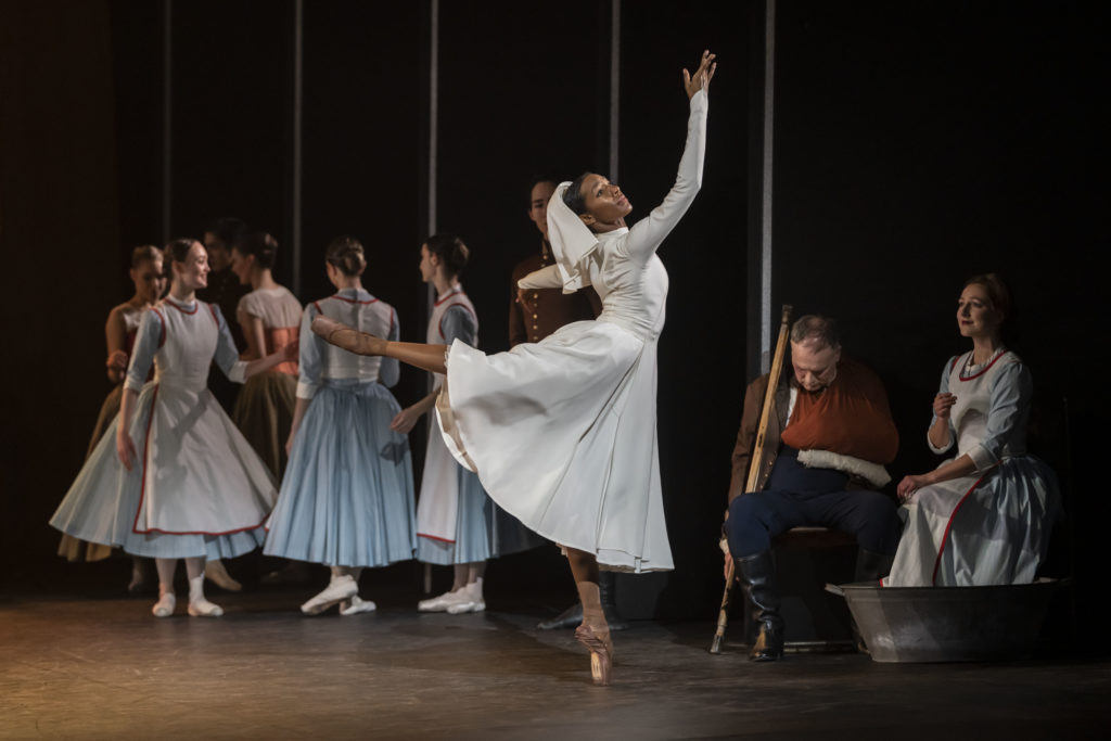 Precious Adams, dressed in a white nurse costume during a ballet performance, does an attitude derriere in effacé with her right leg back. She reaches her right arm up and makes a motion with her hand, while holding her left arm out to the side. She dances close to the wings on stage left; behind her a group of female dancers in blue dresses mill about, while an older man slumps in a chair just off to Adam's left.