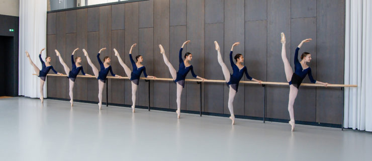 In a large dance studio, seven teenage dance students, all women, pose at the barre aganst the wall in an ecarté derrierre with their right leg up and their left hand on the barre. They all wears a drak blue leotard and short ballet skirt, pink tights and pink pointe shoes.