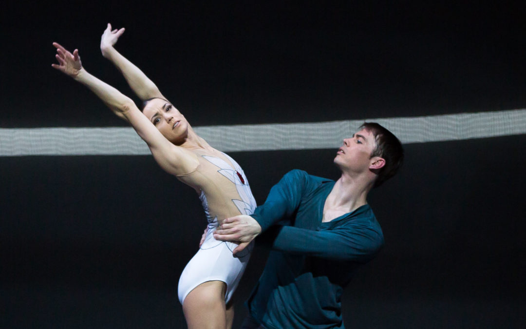 Sophie Martin, wearing a white patterned leotard, stands on pointe in profile with her arms up in a V-shape and arching back slightly, while Christopher Harrison lunges next to her and holds onto her waist. Harrison wears a dark green shirt and pants; both dancers stand in front of a dark backdrop with a white stripe going across it.