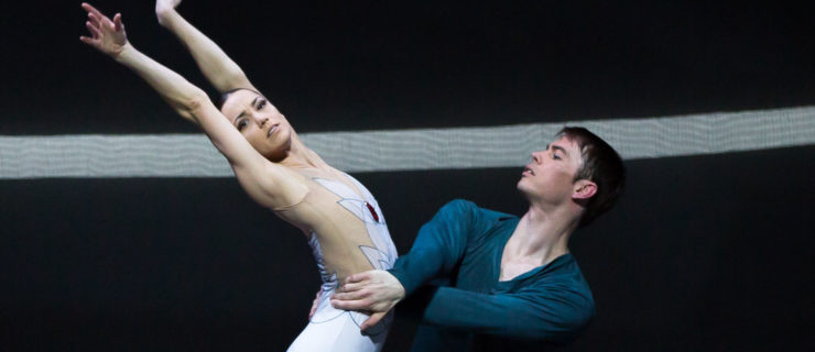 Sophie Martin, wearing a white patterned leotard, stands on pointe in profile with her arms up in a V-shape and arching back slightly, while Christopher Harrison lunges next to her and holds onto her waist. Harrison wears a dark green shirt and pants; both dancers stand in front of a dark backdrop with a white stripe going across it.
