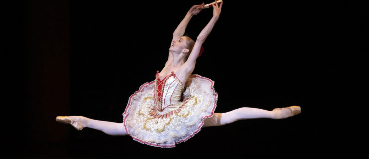 Catherine Hurlin, wearing a white tutu with red and gold trim, flies through the air in a large saut de chat towards stage right. With her arms in high fifth, she holds a closed fan in her right hand and a wears a huge smile on her face.