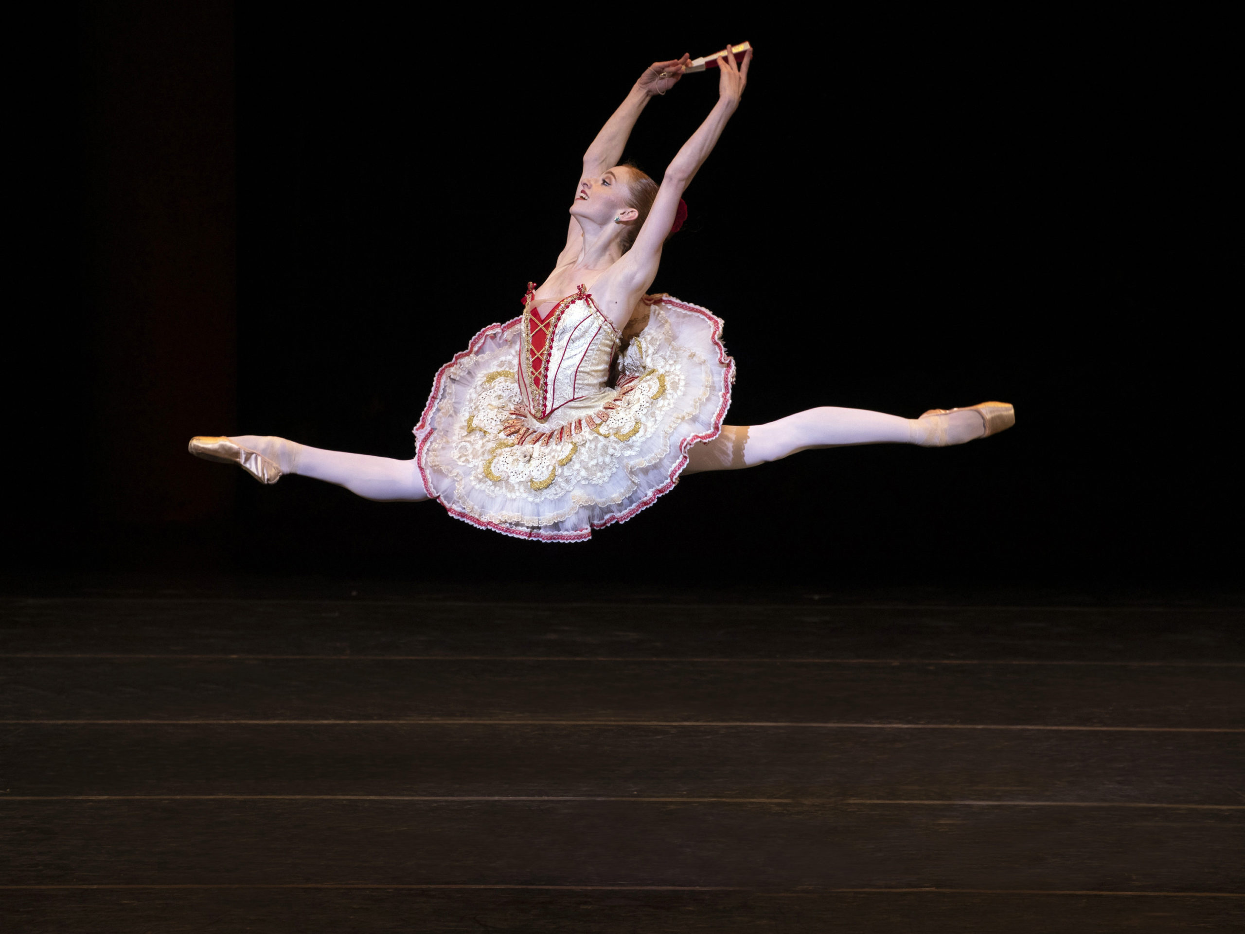 Catherine Hurlin, wearing a white tutu with red and gold trim, flies through the air in a large saut de chat towards stage right. With her arms in high fifth, she holds a closed fan in her right hand and a wears a huge smile on her face.