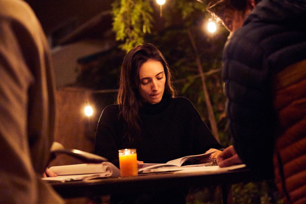 Jahna Frantziskonis, wearing a black turtleneck, sits at a table and reads a script. The outdorr area is lit by a candle on the table and some white lights strung along the trees and fence.