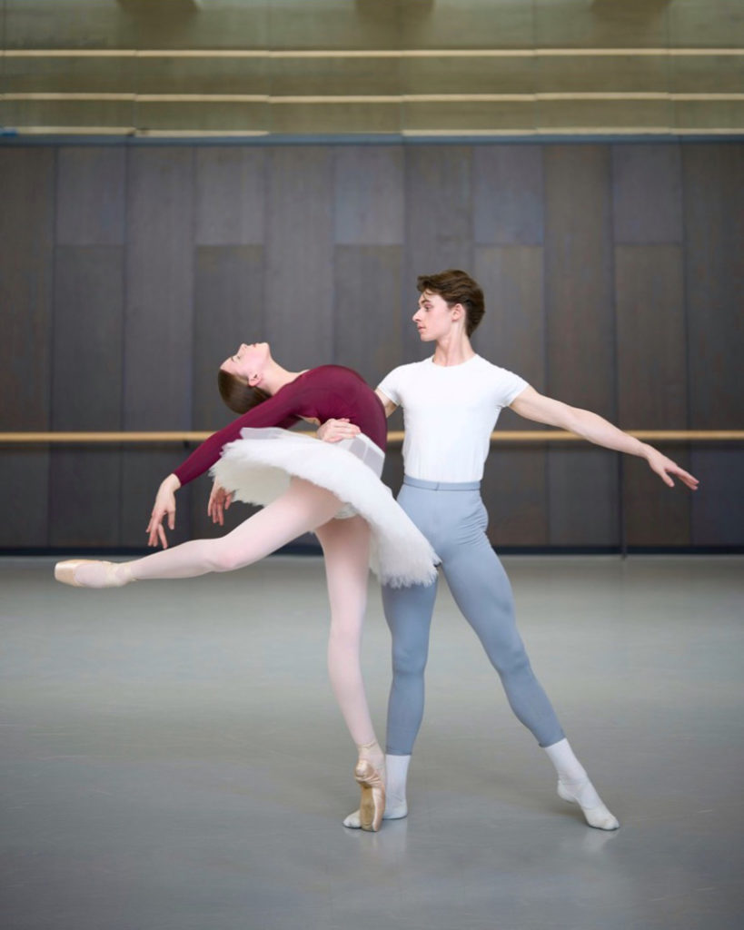 Ava Arbuckle, wearing a burgundy leotard, white practice tutu, pick tights and pointe shoes, poses in a low attitude derriere and does a deep back bend. Mitchell Millhollin, in a white T-shirt, gray tights, white socks and white ballet slippers, holds her by the waist with his right arm and stands on his right leg with his left leg out to the side and his left foot on demi-pointe.