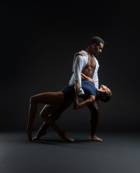 Kyle Davis, wearing an unbuttoned white shirt, brown tights and ballet slippers, lunges onto his left leg and holds Gabrielle Salvatto around the ribcage as she leans back, crouching on pointe. Salvatto wears a blue high-neck leotard and holds onto Davis' right arm and shoulder with her hands. They look at each other.