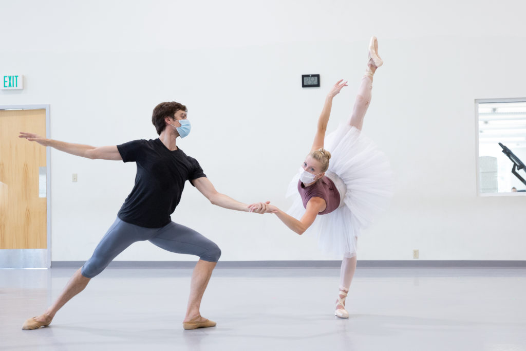 Steven Loch and Dawn Atkins rehearse a ballet in a large rehearsal studio with white walls. Loch lunges on his left leg and holds Atkins' left hand with his right as she lowers into a croisé penché arabesque with her right leg back. Loch wears knee-length blue tights, a black T-shirt and tan ballet slippers, and Atkins wears a purple leotard, white practice tutu, pink tights and pink pointe shoes. They both wear face masks.