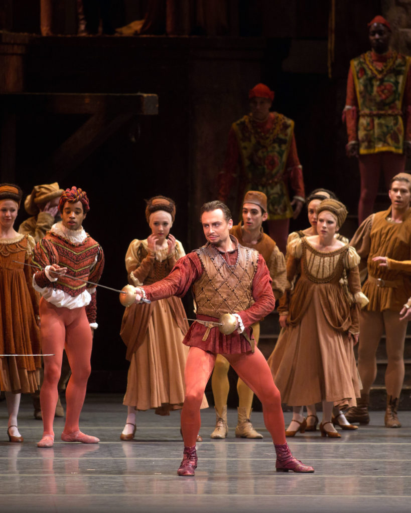 Roman Zurbin stands with his legs slightly apart and bent, and holds a sword in both hands during a fight scene in the ballet "Romeo and Juliet." He wears a costume of reddish brown tunic with a brown overlay, orange tights and red boots. Behind him, townspeople in various medieval costumes look on fearfully.