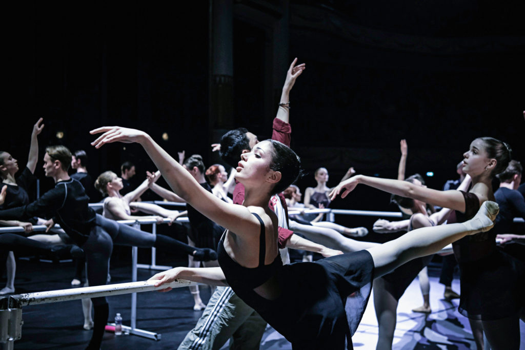 A dancer is shown performing a second arabesque on a portable barre, raising her right leg.  She is surrounded by other dancers doing the same step on several portable ballet bars lined up on stage.  She wears a black leotard, black skirt, pink tights and ballet shoes.