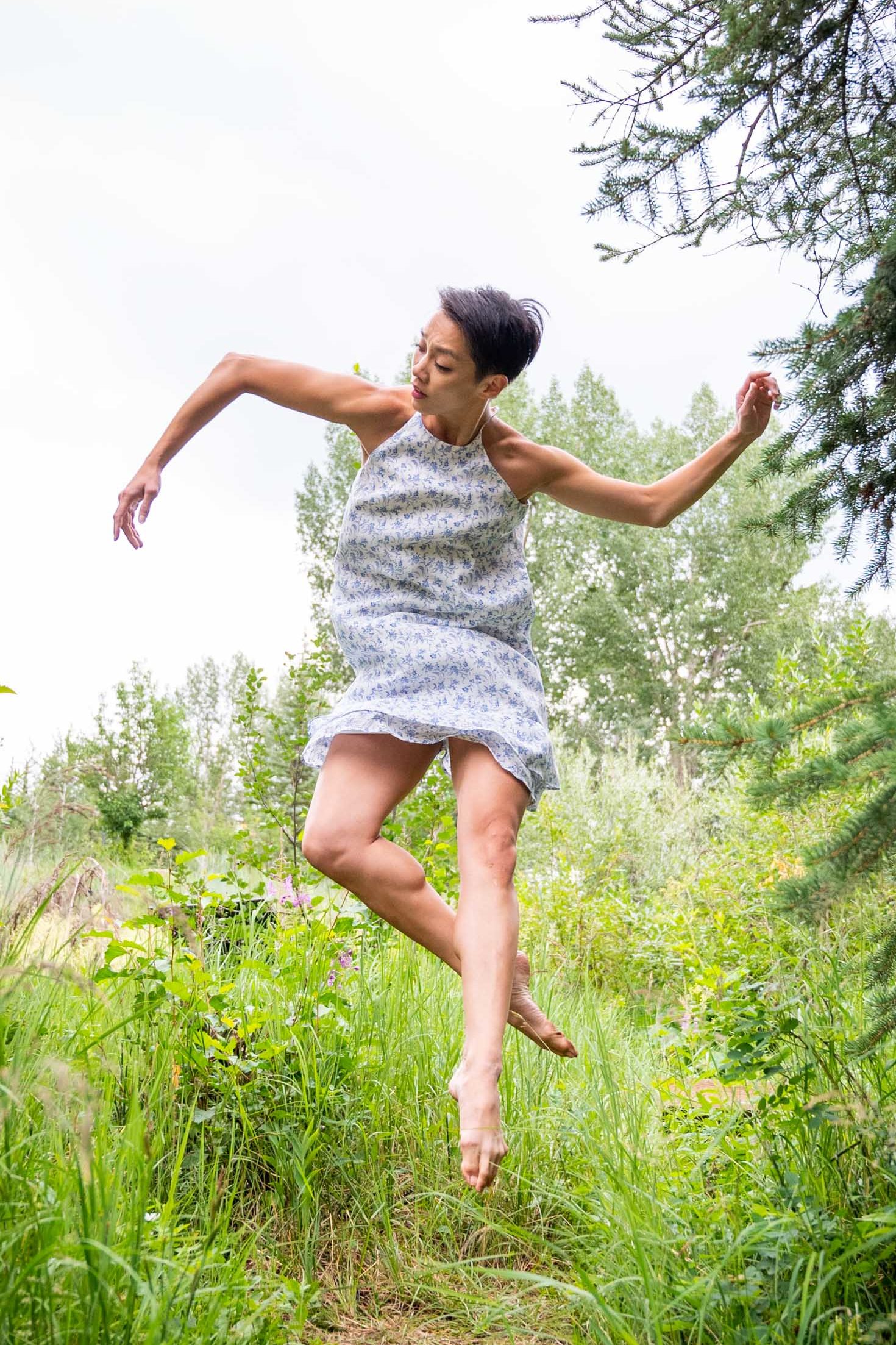 Caili Quan, with her short dark hair down, jumps in a low contemporary coupé derriere in a flowy white sheath dress. Her arms make an "S" shape, her right arm bent at the elbow to point down and her left pointing up. She looks slightly down on the right diagonal, past her arm. She dances in light green grass and brown dirt, with trees beside and behind her, the sky a light white-ish gray behind her. 