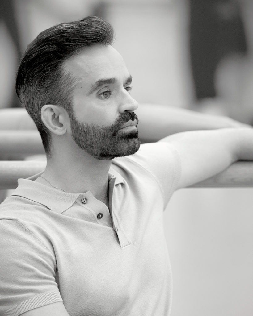 Black and white image. Aaron Watkin, shown sitting chest-up, rests his left arm on a ballet barre against the mirror as he watches rehearsal in a studio. He has dark short hair and a clean-cut short beard, and he wears a light-colored polo shirt.