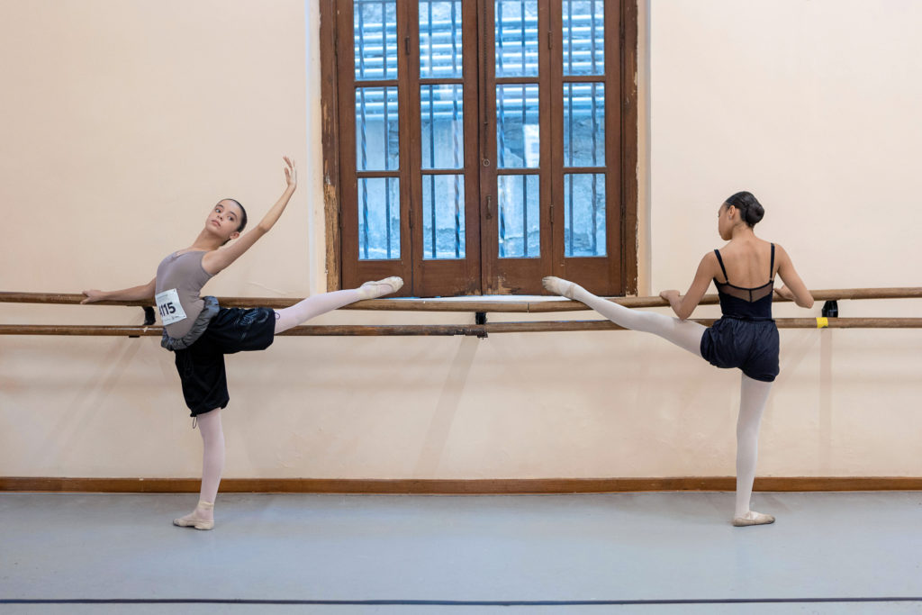 Two young female dancers warm up in a studio before an audition class. The girl on the left, in a light brown-gray camisole leotard, wears black garbage bag warmup shorts over her pink tights and ballet slippers. She rests her left leg in an arabesque stretch on the barre, facing outward and in a combré back with her right arm on the barre and her left arm overhead in 5th en haut. She wears a paper number safety pinned to her leotard and looks out past her working elbow into the studio. The dancer on the right rests her left leg on the barre in an a la seconde stretch, facing in toward the barre, and looks at her pointed foot. A large wooden-frame window divides the cream-colored wall between them.