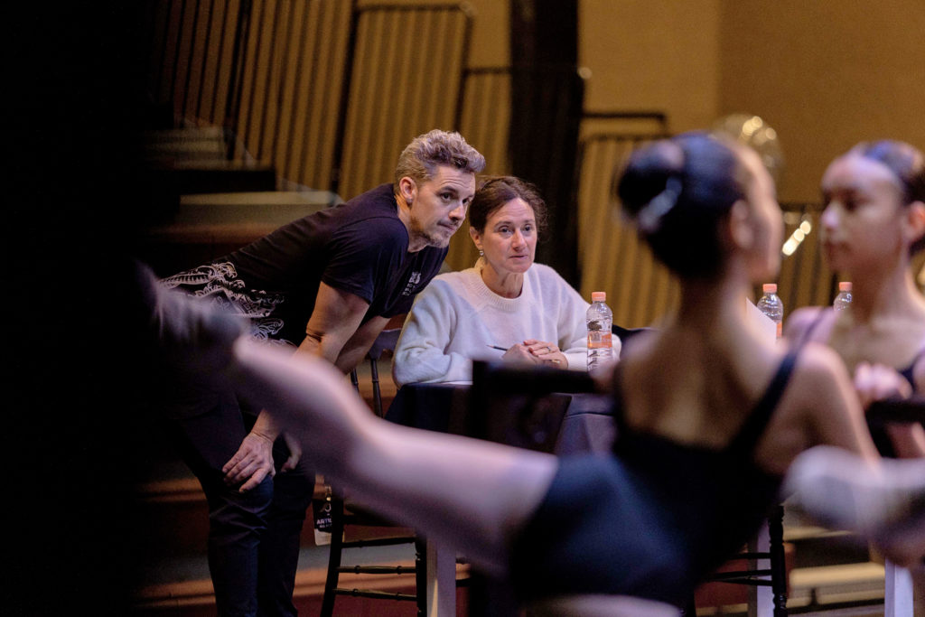 In the blurred foreground, two young female dancers execute an arabesque with their hands on the barre, facing in toward each other. The focused background shows a male and female who lead the audition class in front of risers. The woman, with her brown hair up and wearing a white sweatshirt, sits at a desk with water bottles and papers, holding her pen in her folded hands. The man, in a black t-shirt, black pants and a knit wrap around his mid-section, leans toward her with his arms resting on his bent knees. They both watch the dancers eagerly.