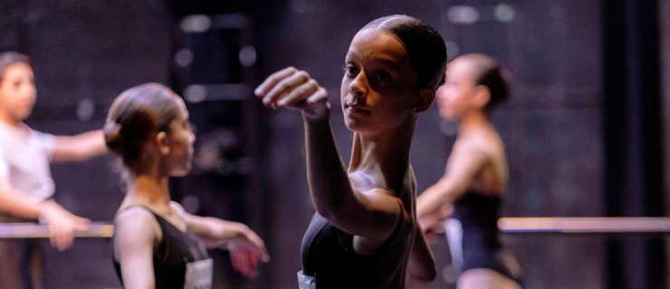 A young female dancer, shown hips-up, stands sideways at the barre onstage for an audition. She wears a black camisole leotard and pink ballet tights, and her dark hair is slicked back into a low bun with a middle part. She extends her left arm in allongé out from the barre, looking out past her fingertips with a slight smile, eyebrows raised gently in concentration. She wears a paper number safety-pinned to the front of her leotard. Two other girls, dressed similarly, and a young male dancer in a white t-shirt, black tights, and white socks,complete the same motion in the background.