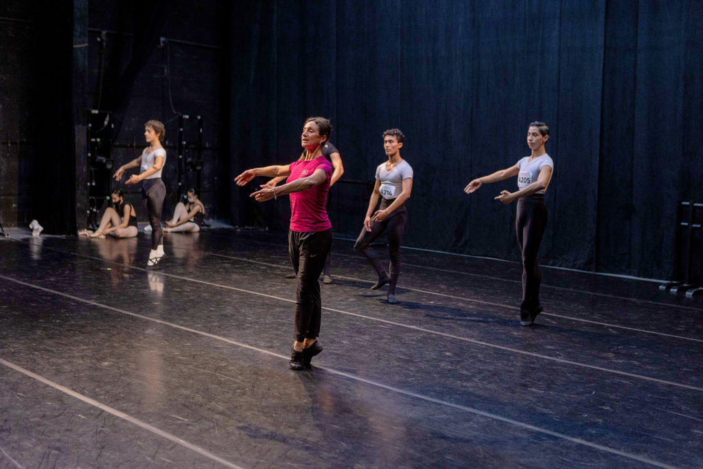 A woman in a hot pink t-shirt, black athletic pants and black teaching shoes leads class onstage to four young male dancers. She stands in sous-sus with her arms in 3rd position, preparing for a tour en l'air. The dancers behind her mimic her movements, dressed in white t-shirts, black tights and shoes. They wear paper numbers, safety pinned to their fronts, for the audition class.
