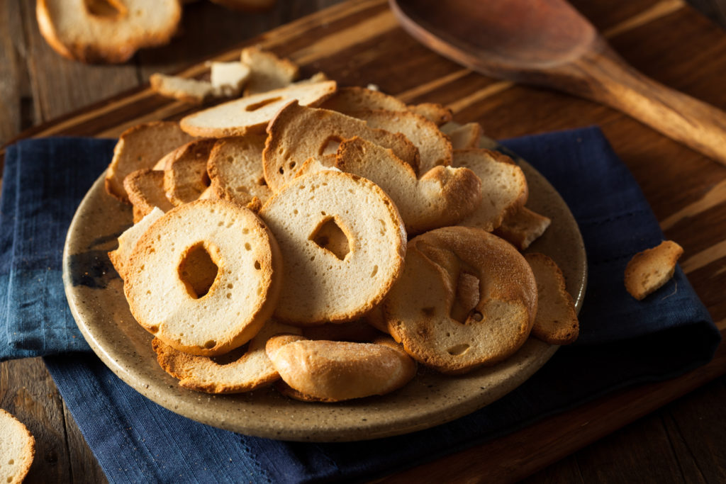 A pile of bagel chips sits on a light brown ceramic plate. The plate is on a folded blue linen napkin on a wooden table.