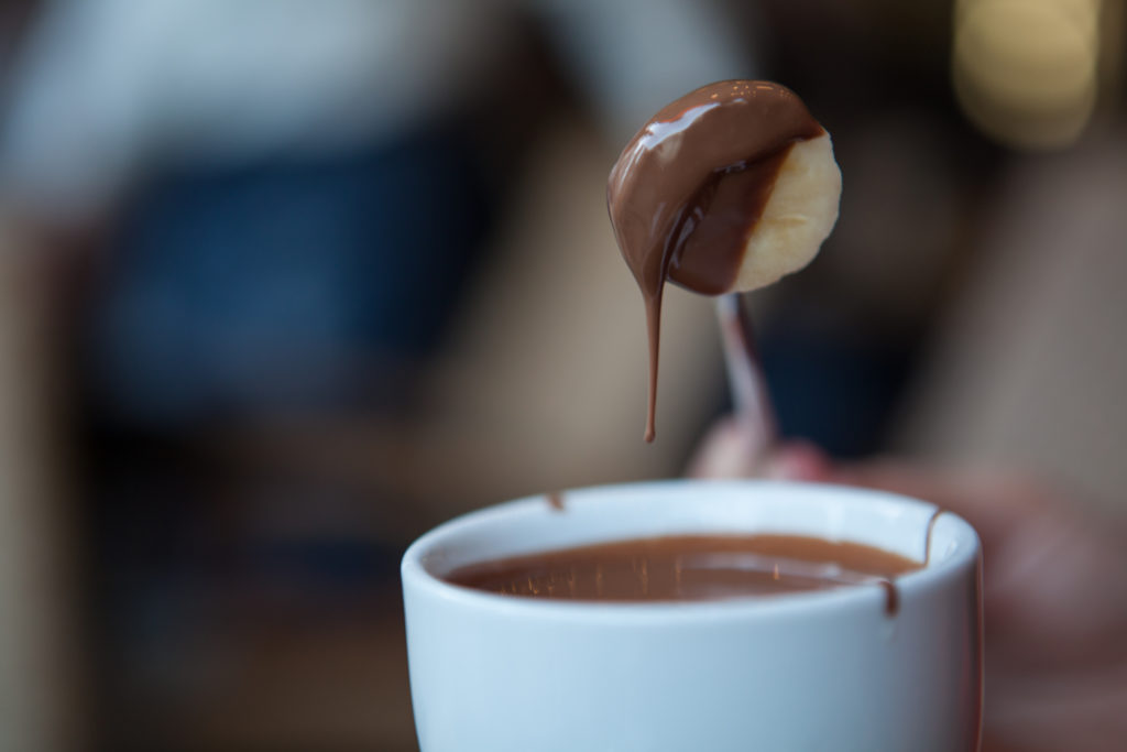 A white cup of melted chocolate is in front of a blurry background. A metal skewer with a banana slice half dipped in warm chocolate is being lifted from the cup.
