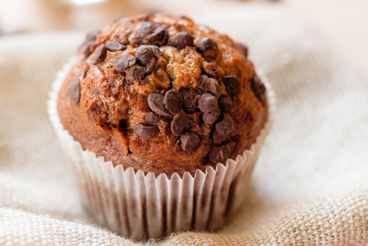 An orange-colored muffin with a white wrapper is stuffed with chocolate chips. It sits on a beige towel.