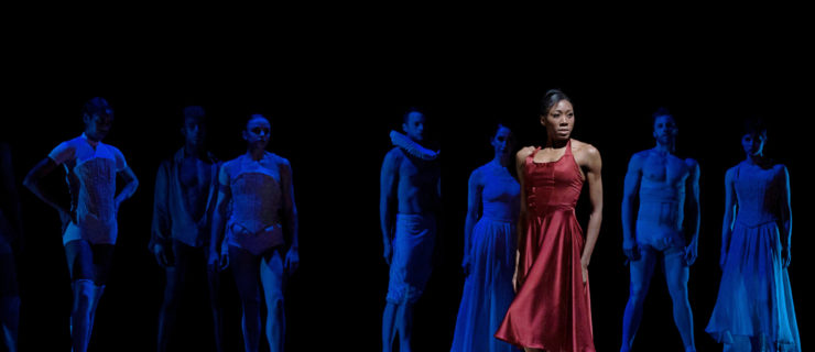 Claudia Monja, wearing a red silk dress and flesh-colored pointe shoes, and with her black hair in a French twist, stands with strength in a spotlight onstage. Her body is turned toward the left, but she turns her torso slightly as she looks straight at the audience with an intense but pensive expression, her energized arms resting at her sides. Her left leg bevels on pointe turned in. The ensemble behind her, dressed in modernized and minimalist Shakespearean garb, stands watching her in dark blue lighting.