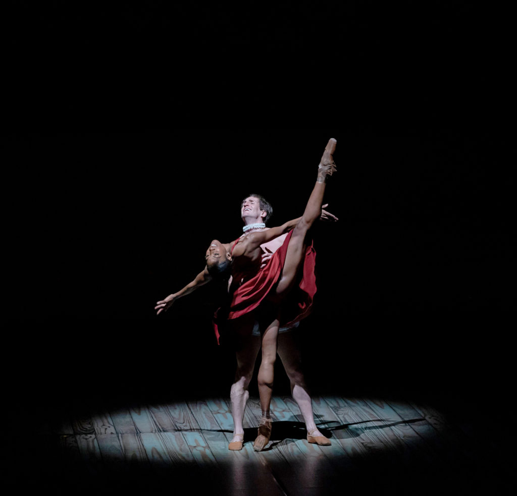 Claudia Monja, in a red silk dress and flesh-tone pointe shoes, stands on pointe on her left leg with her right leg in a la seconde, vertical to the stage. She leans back and looks at the audience with emotion, arms spread wide with palms up. Nicolas Scheuer holds her by the waist, standing in parallel behind her, his face full of emotion as he closes his eyes and tilts his head up with his mouth open. He wears nude ballet flats, white bloomer shorts and a white ruffle collar. They dance in a spotlight on a pitch black stage, the floor covered in a wooden pattern.