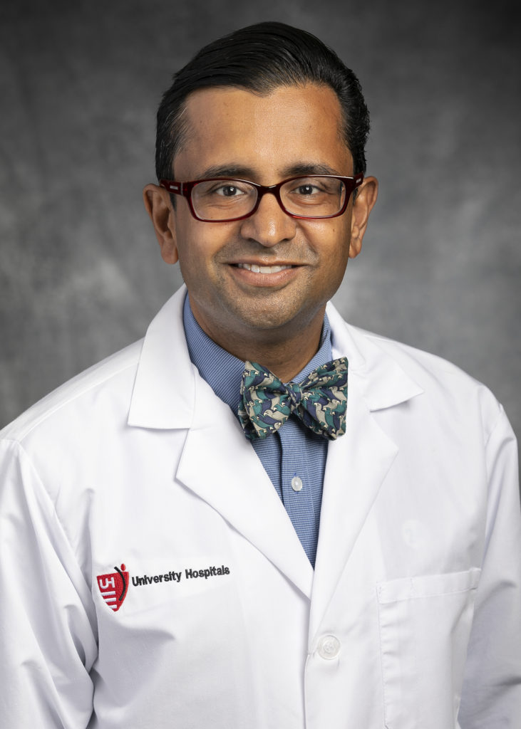 A smiling male Indian doctor with glasses, dark hair, a patterned bowtie and a white medical coat.