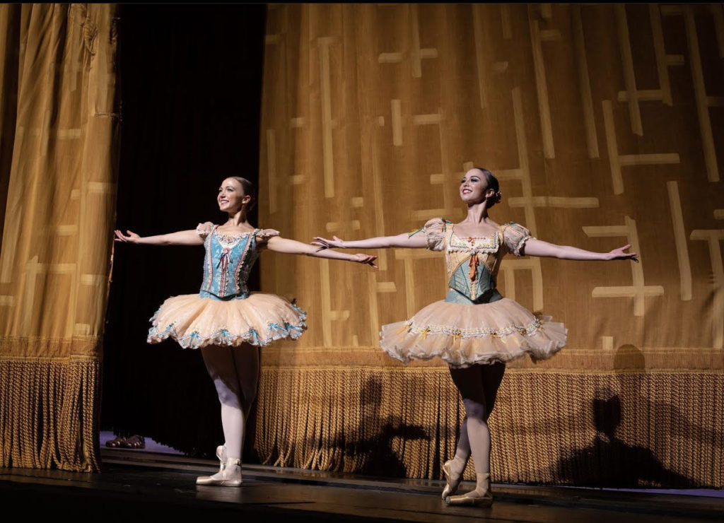 Paulina Waski and Katherine Williams bow in front of a golden curtain onstage. They wear butter yellow ruffly tutus with sage green, light blue, and yellow short-sleeved corset bodices with a pink ribbon at the front and white and pink lace details. They wear pink ballet tights and pointe shoes with their dark hair slicked back into a low bun. They stand in B plus on their right legs, arms outstretched and upturned in a generous welcoming posture, smiling out to the audience proudly with chests lifted.