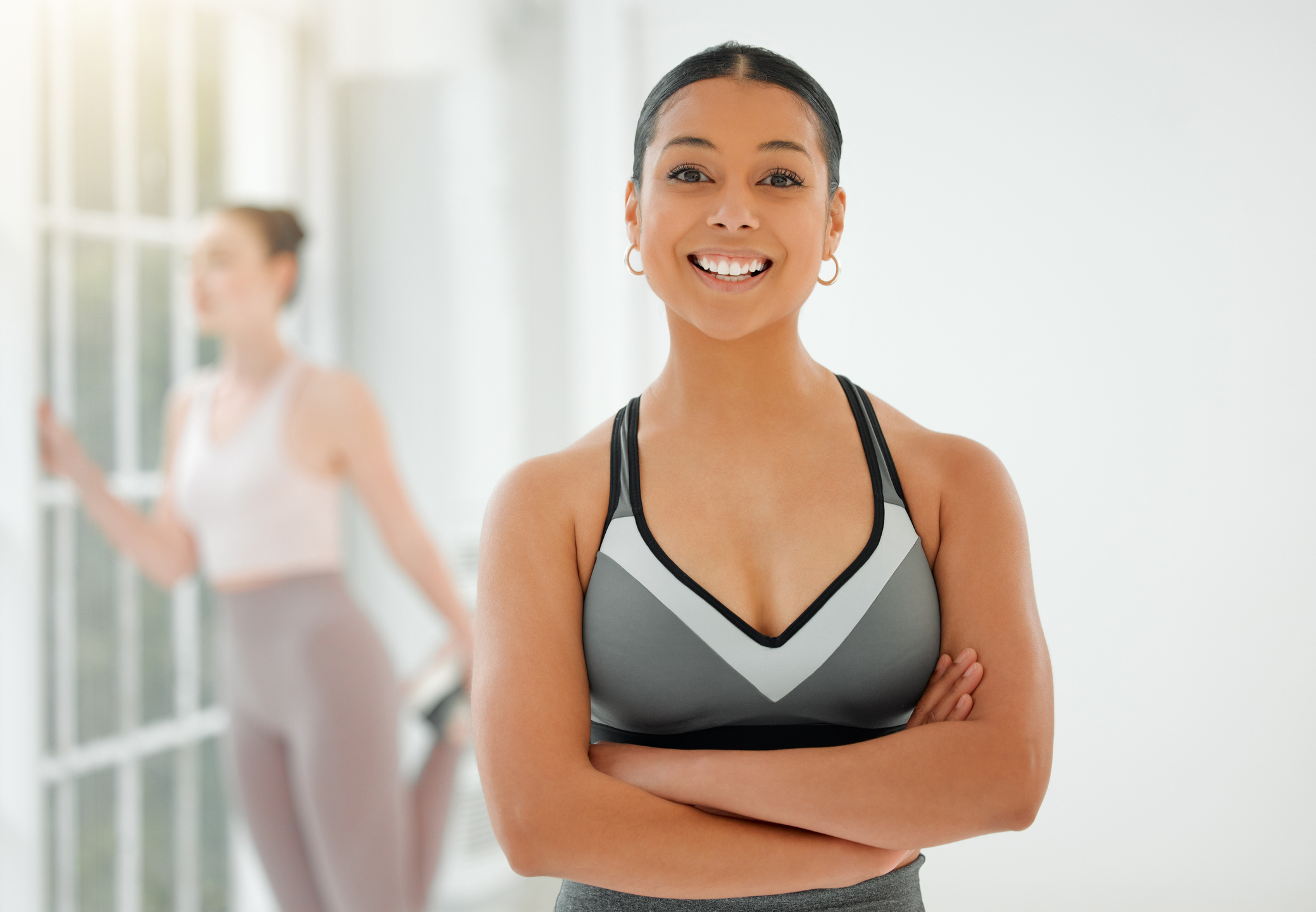 A young woman in a gray halter-top leotard is shown from the waist up in a brightly lit studio. She smiles widely at the camera and crosses her arms. Another woman in workout clothes stretches her left quadriceps and holds onto the window for support.