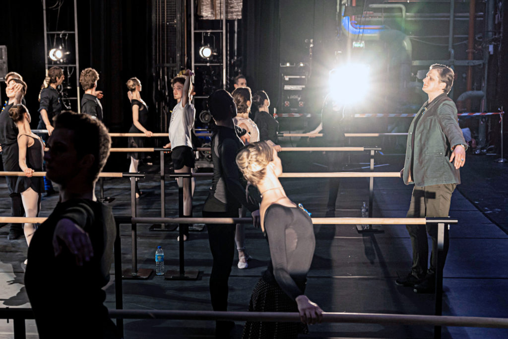 A male ballet instructor stands towards the front of a stage and teaches a class full of professional ballet dancers, who stand at rows of ballet barres. The stage is dimly lit from the side with work lights.