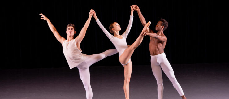 Three ballet students dressed in white dancewear pose to make a trio formation. A female student stands in the middle on pointe with her right leg in developpé croisé devant. She holds the hands of two male dancers on either side of her; the one on the left poses in arabesque with his left leg up, and the one on the right poses in tendu derriere and holds her right ankle with his left hand.