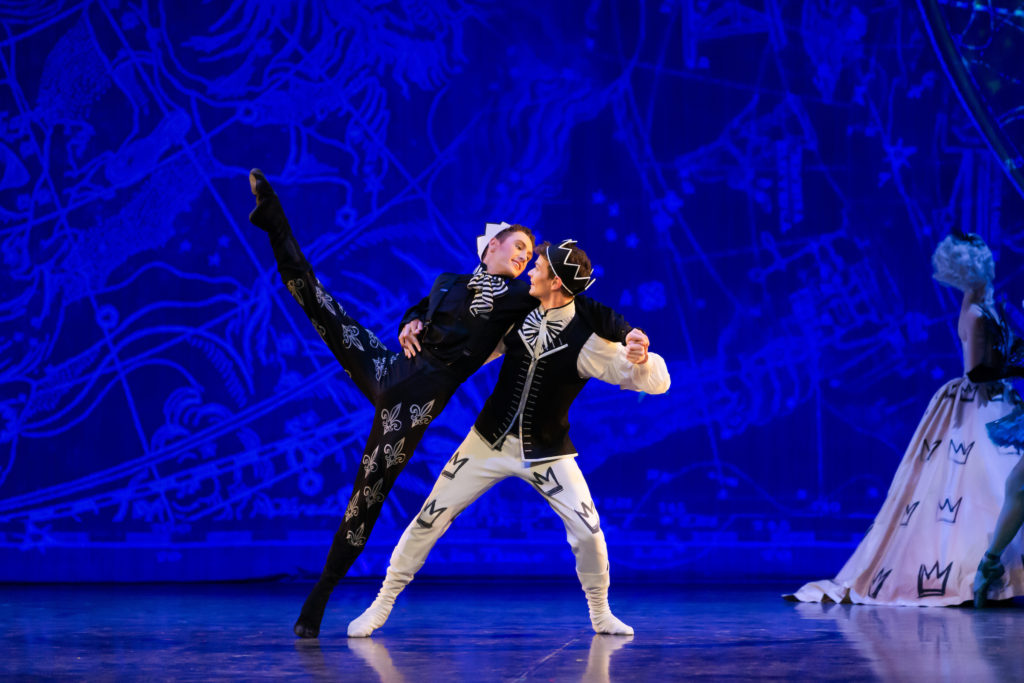 Joshua Guillemot-Rodgerson, as Prince Charming, and Shae Berney, as Prince Dashing, dance together in a pas de deux onstage in front of a deep blue textured backdrop. Guillemot-Rodgerson wears white tights with black crowns down the sides, a black waist jacket, flowy white peasant blouse, a black and white striped bow tie, white dance boots and a black fabric crown with white trim. Berney wears black tights with white fleurs de lis, a black jacket, black dance boots, black and white neck scarf, and white fabric crown. They gaze into each others' eyes happily, Berney in battement a la seconde with his left leg, his left side leaning on Guillemot-Rodgerson for support. G-R, in a slight lunge, extends his left arm to the side and bends his elbow to hold Berney's hand as he reaches his left arm behind G-R's shoulders. A woman in a large white dress with black crowns, with a large white royal wig, faces her back to them in the right corner of the photo.