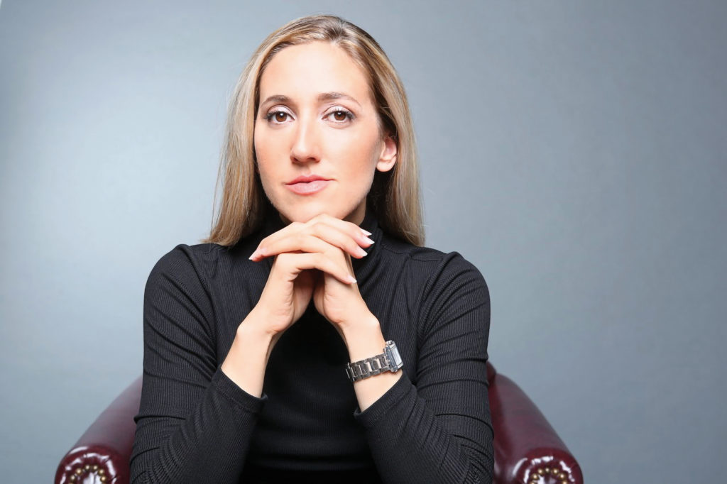 Melissa Barak, a young professional woman with long blonde hair, is shown sitting chest-up in a long-sleeved black shirt on a comfortable brown leather chair in front of a light gray background. She wears a silver watch on her left wrist as she folds her hands together, resting her chin on tip of them as she looks confidently into the camera with a slight smile.
