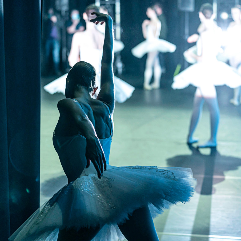 A female dancer in a white tutu practices a tendu croisé derriere in plié backstage, her arms extended in a long first arabesque line and her head angled towards the floor. Meanwhile, dancers in white tutus practice in the background onstage.
