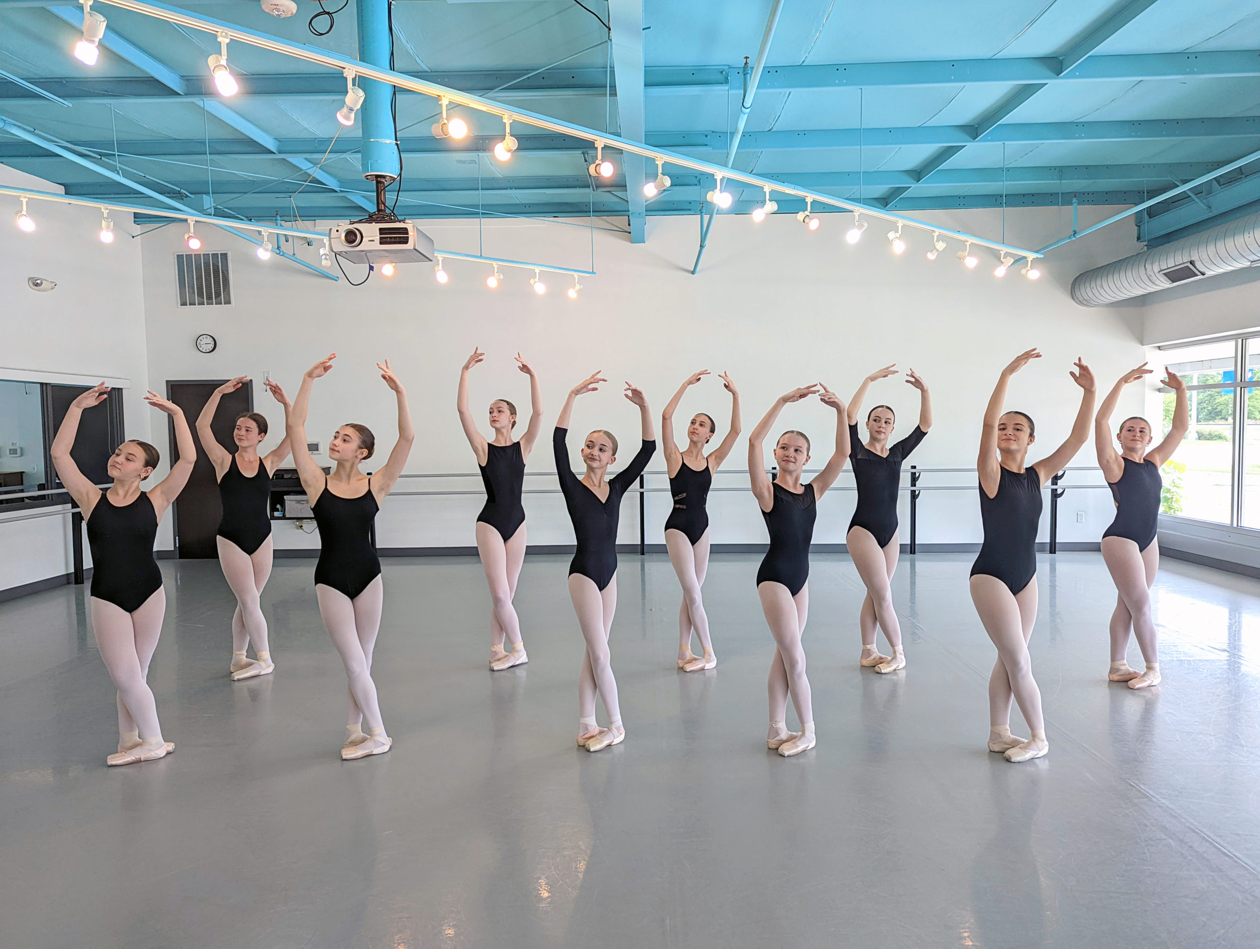 A group of 10 female students in black leotards, pink tights, and pink pointe shoes stand in two staggered lines of five in a sunny dance studio. They all pose in fifth position with their right foot in front and their arms in high fifth position, looking slightly to the right.