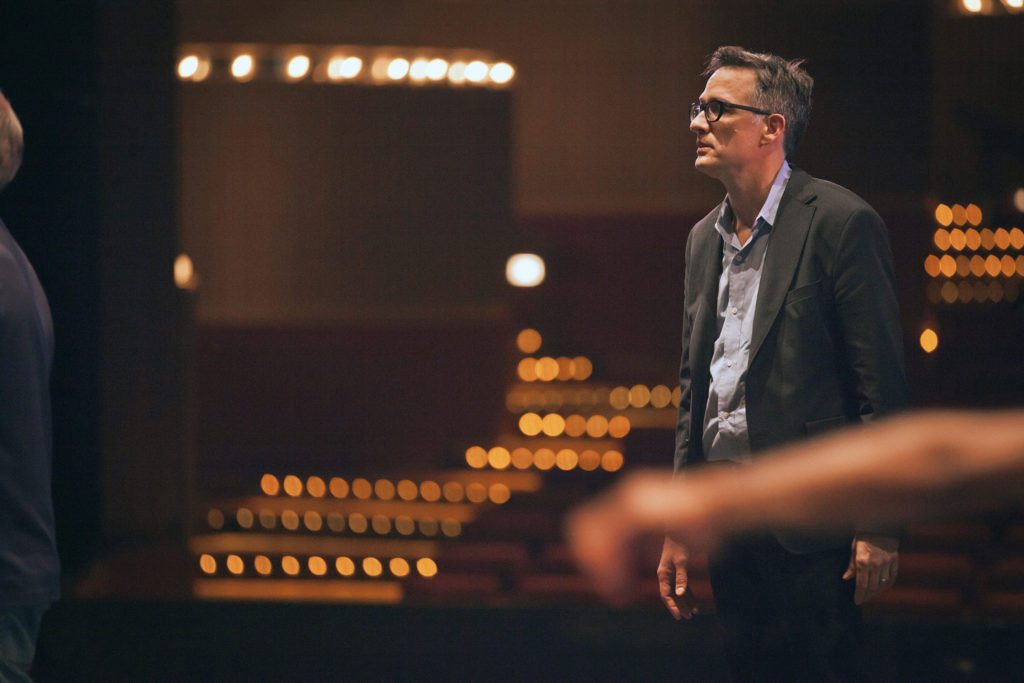 Paul Vasterling, at the far right, stands in the midground in a darkly lit theater space wearing a gray suit and light blue button up. He wears glasses and faces the stage (out of screen), watching intently with his arms at his sides. The house lights in the theater are a soft orange yellow color.