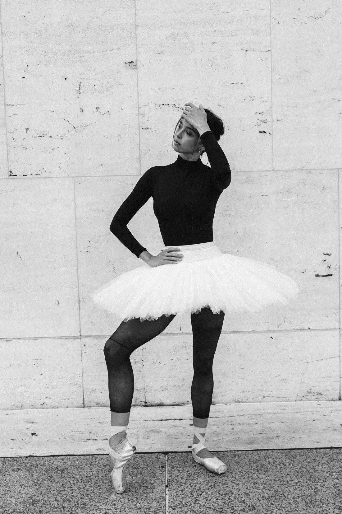 Paulina Waski stands casually outside the Met Theatre in a black and white photograph. She wears a black long-sleeved leotard and cut-off tights, a white practice tutu and pointe shoes. Her dark hair is in a loose bun, and she stands with her right leg slightly propped up on pointe. Her right hand rests on her hip, her left bent up to brush hair away from her face. She looks out contemplatively to the right in front of the white marble wall..