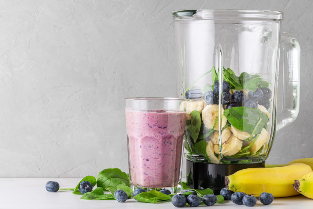 A clear glass with a purple smoothie sits next to a blender filled with banana slices, blueberries, and spinach. Two unpeeled bananas, spinach leaves, and several blueberries are scattered at the base of the glass and blender.