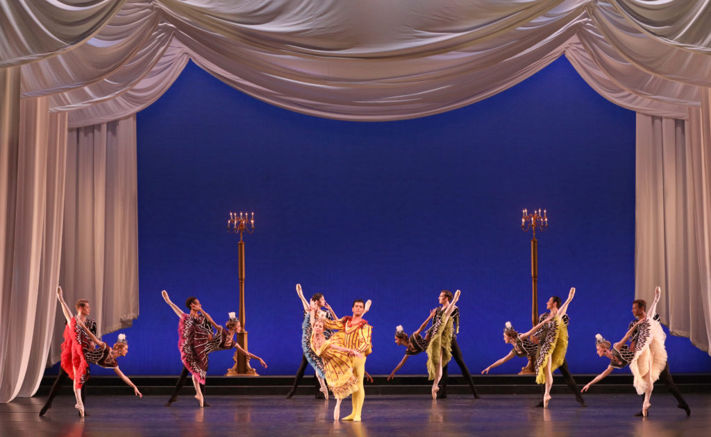 Seven pairs of partners dance onstage in front of a deep blue backdrop and swooping golden curtains that line the top and sides of the stage. The women wear various shades in adorned dresses, with stripes, dots, and triangles, as well as pink ballet tights and pointe shoes and matching head pieces with tall white flowers. Six of the pairs pose in a partnered penché, all facing toward the middle of the stage. The center couple, in yellow, poses in a partnered croisé arabesque in slight penché, her leg extending behind his head as she allongés her right arm on the high diagonal and her left on the low so her partner can hold on to her forearms. They look out toward the audience confidently. 