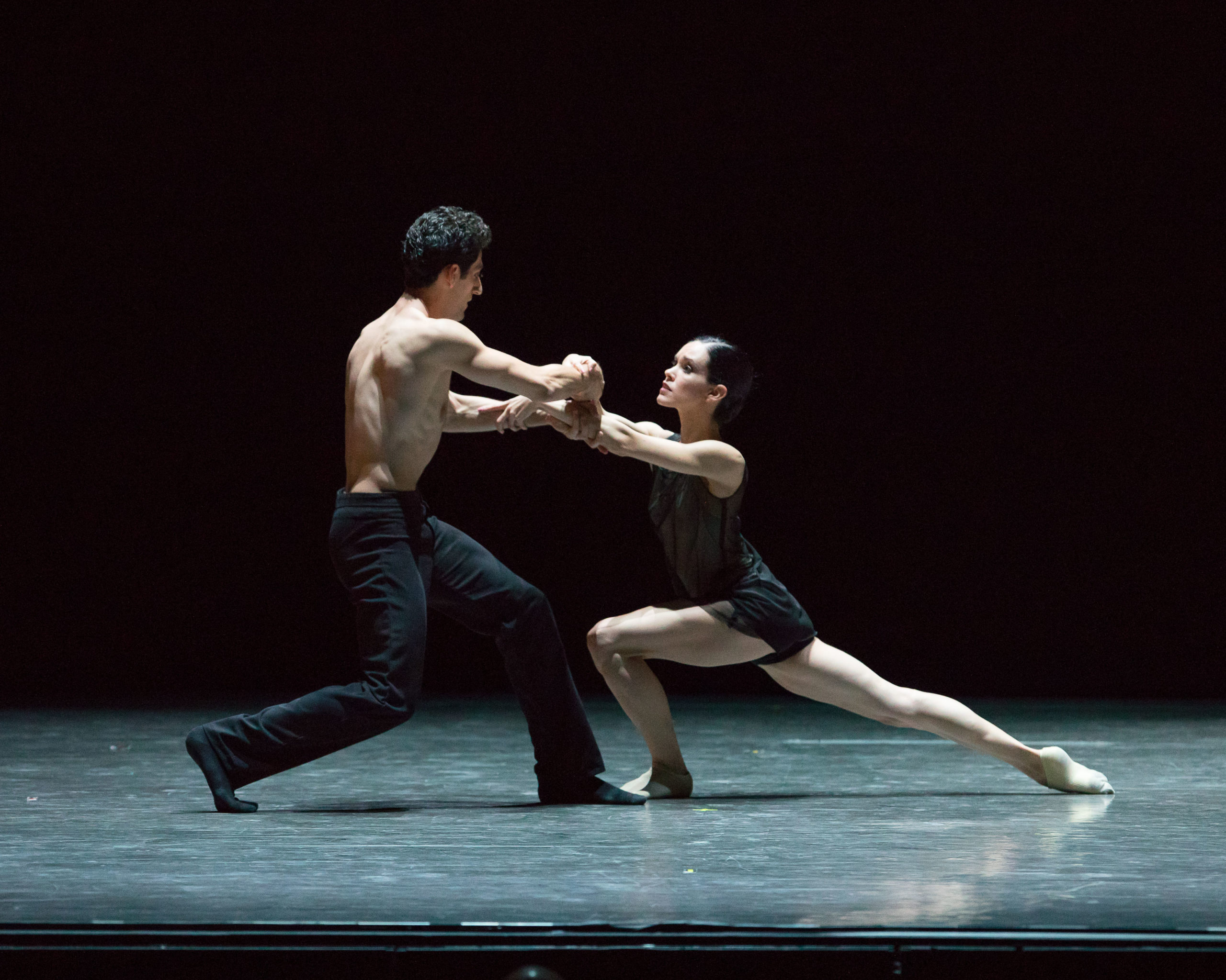 Tigran Sargsyan and Petra Conti firmly grasp each other's forearms, arms crossed as they gaze into each other's eyes. Conti is in a low lunge, her right leg extended back with pointed toes.