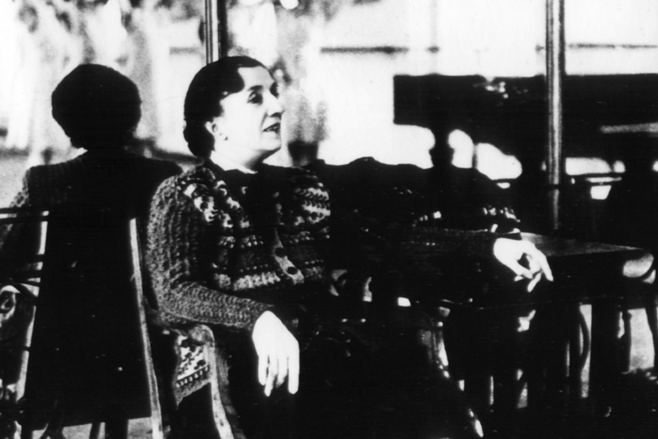In this black and white archival photo, Agrippina Vaganova sits in a chair at the front of the studio, her back towards a mirror, and watches dancers (unseen) in class. She wears a wool sweater cardigan and long dark skirt. A grand piano is seen in the mirror's reflection.