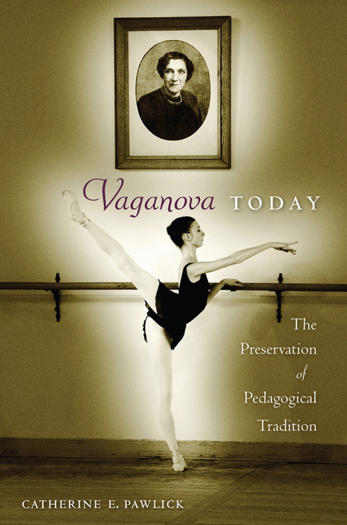 The cover of a book titled "Vaganova Today: The Preservation of Pedagogical Tradition" shows a sepia-toned photo of a female teenage ballet student practicing a high second arabesque at the barre with her right leg raised. The dancer wears a black leotard and short black ballet skirt, pink tights and ballet slippers with ribbons attached. Above her, a portrait of Agrippina Vaganova hangs on the studio wall.