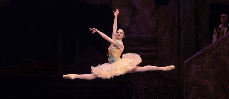 Paulina Waski grins as she saut de chats in "Don Quixote" onstage. She wears a butter yellow ruffly tutu with a sage green and yellow short-sleeved corset bodice with a pink ribbon at the front and white lace details. She wears pink ballet tights and pointe shoes with her dark hair slicked back into a low bun. Her arms reach energetically in the 3rd arabesque line as she smiles toward the audience.
